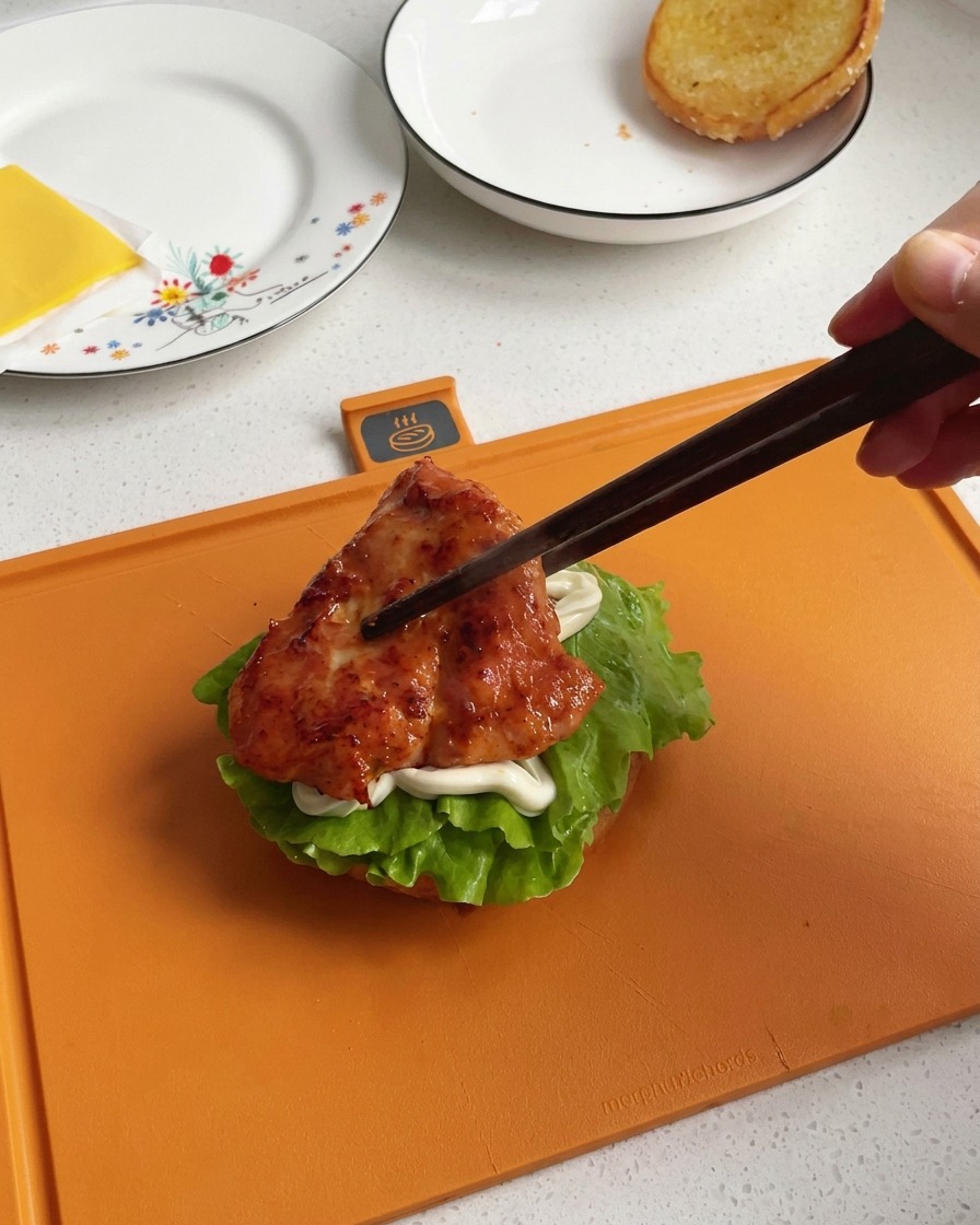 濃い色の木箸を使い、ツヤよく香ばしく焼き上がったチキンをバーガーの層の上に移している手。