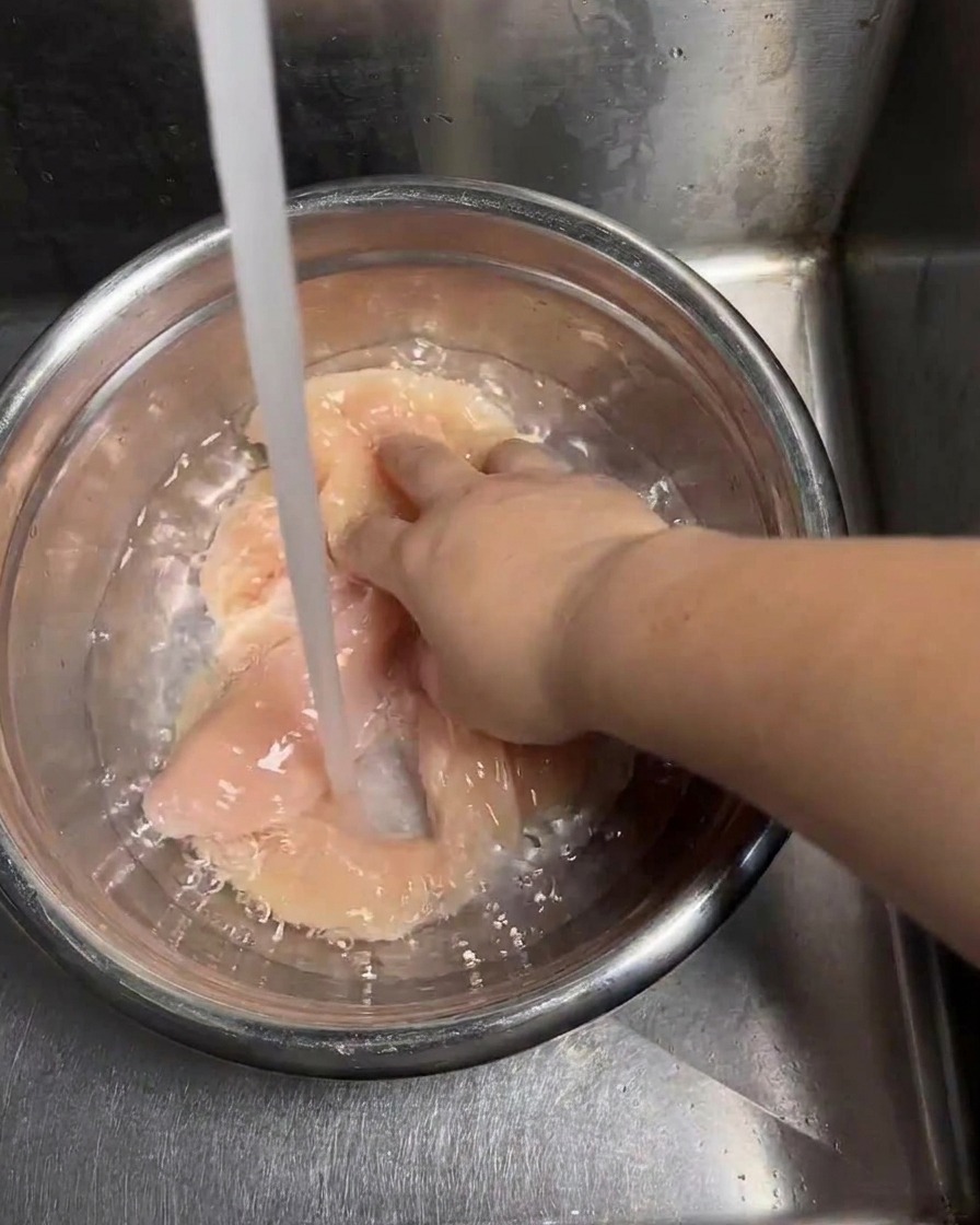 生のチキンカツが入ったステンレスボウルに冷たい水道水を注ぎ、手で肉を動かしながらきれいに洗い流している様子。