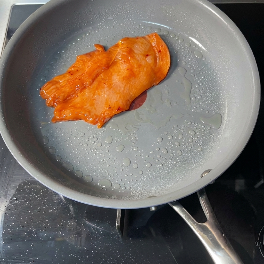 表面にうっすらと油が浮いたグレーのノンスティックフライパンの中で、オレンジ色の味付けされたチキンカツが1枚焼かれている様子。