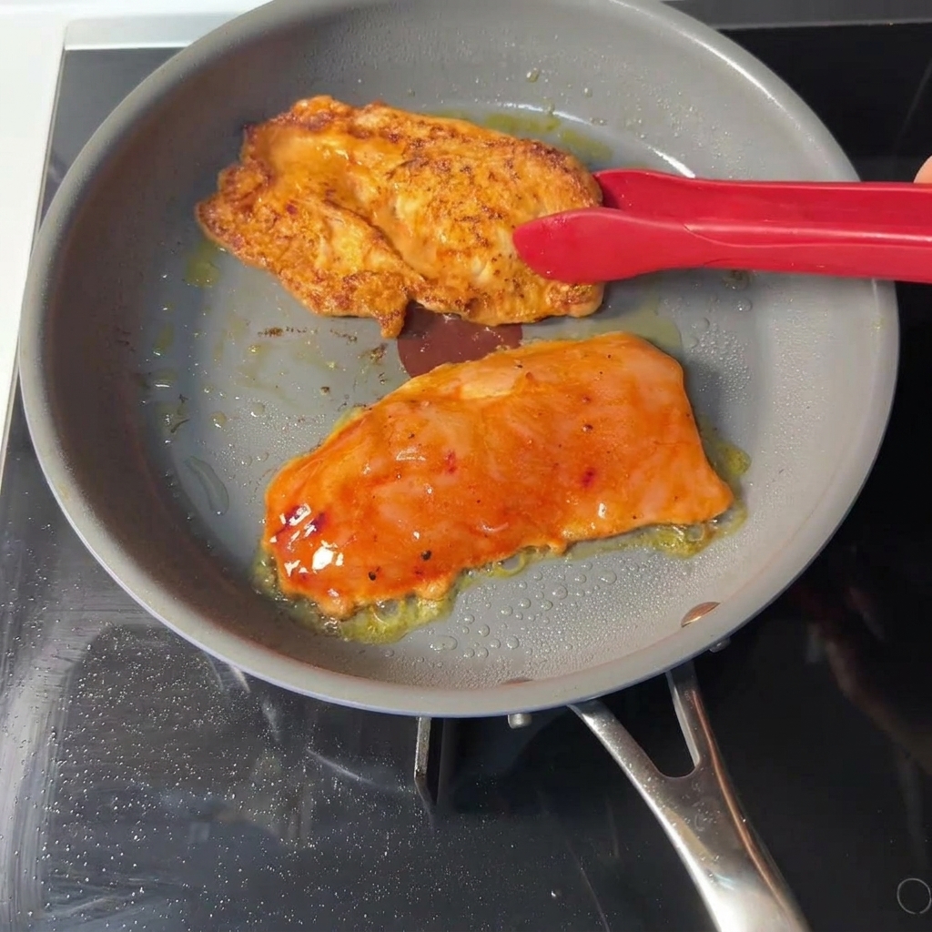 グレーのフライパンの中で、赤いトングを使ってオレンジ色のマリネされたチキンカツをひっくり返し、焼けた面にきれいなきつね色の焼き目がついているのが見える様子。