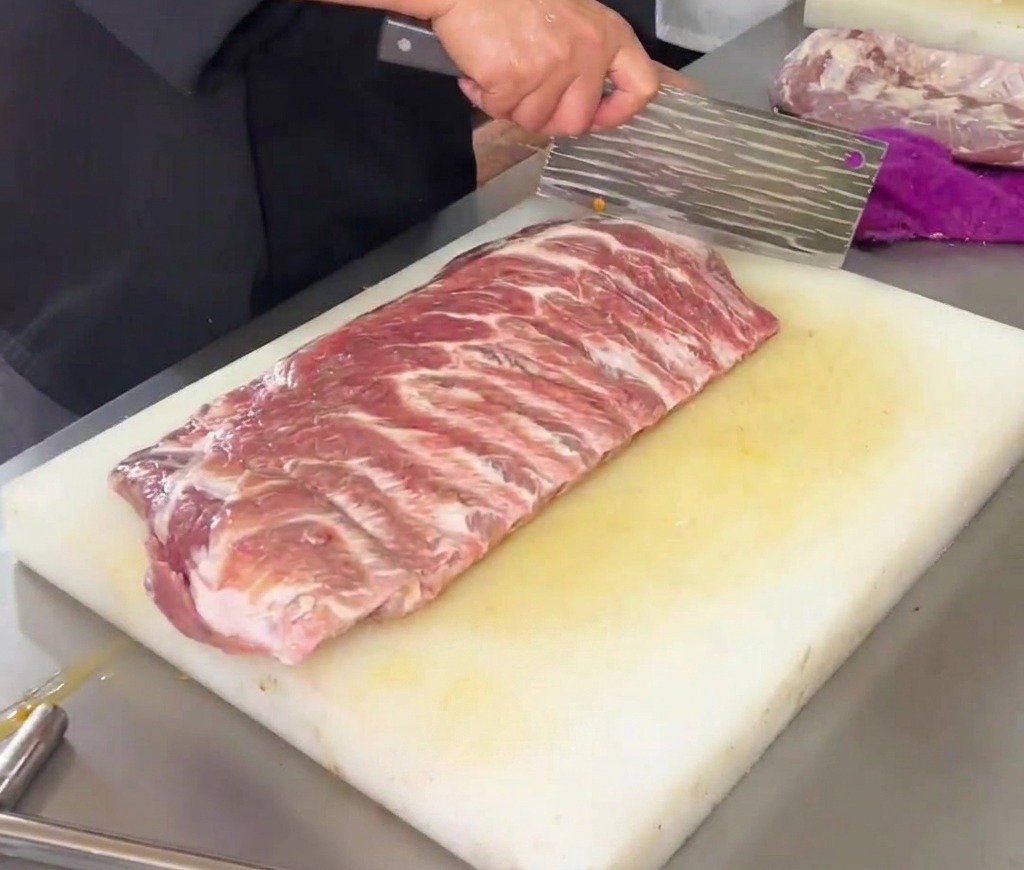 白いプラスチック製まな板の上で、大きな中華包丁を使って生の豚リブ肉にダイヤモンド状の切れ目を入れるシェフの手元。