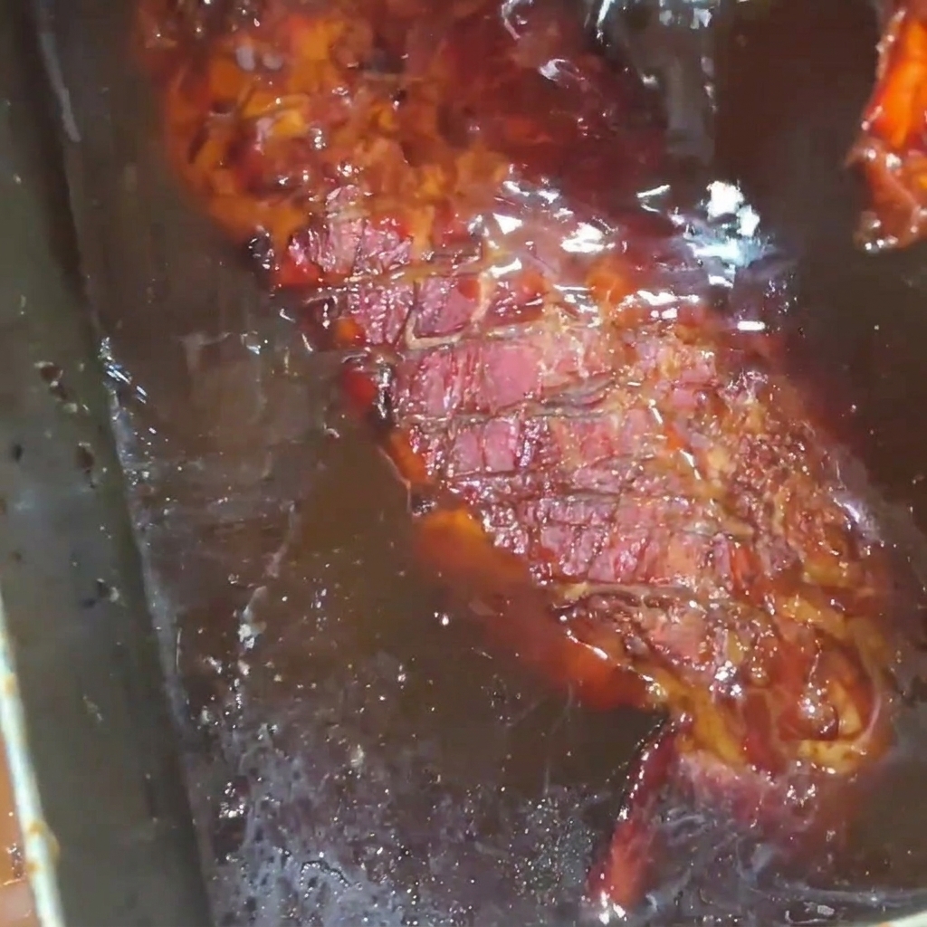 ステンレス製のトレイの中で、こんがりと焼き上がったポークリブに、艶やかで濃厚なハニーシュガーシロップのグレイズをたっぷりと回しかけている様子。
