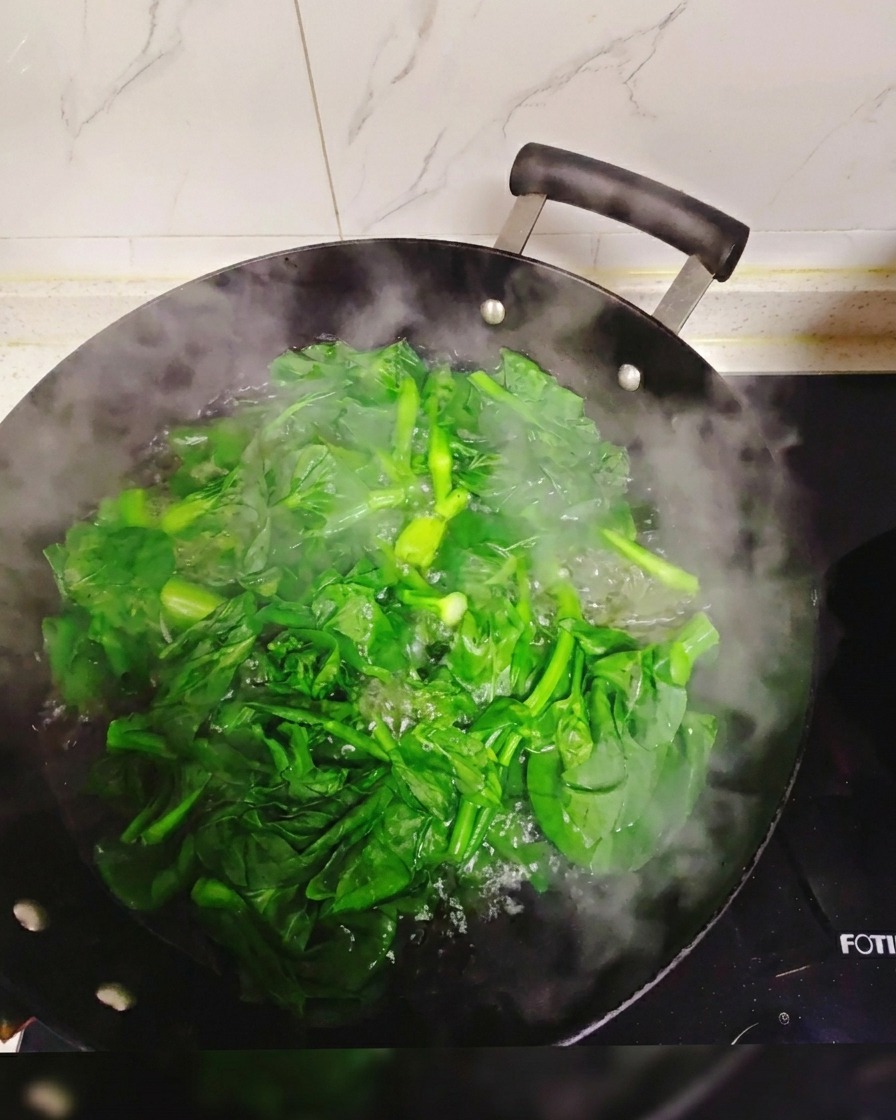 大きな中華鍋にたっぷりと沸騰したお湯の中で、鮮やかな緑色の青菜をサッと茹でている様子。