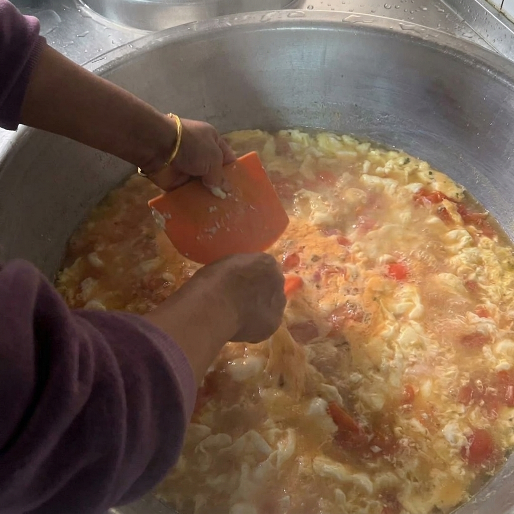 沸騰したトマトスープが満ちた大きな鍋に向かって、オレンジ色のプラスチックスクレーパーを使い、柔らかい生地を小さく削ぎ落としている様子。