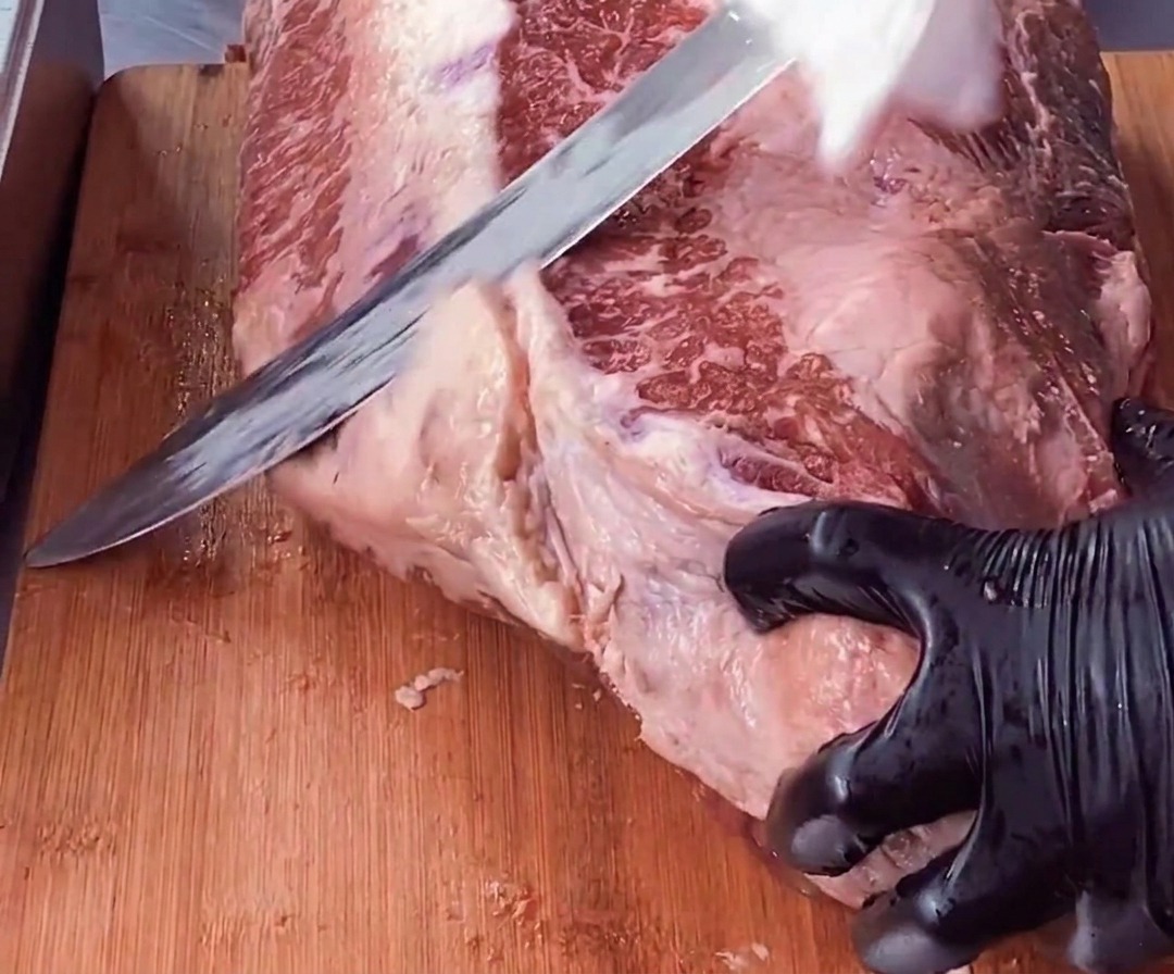 手袋をはめた手でブリスケットの塊を固定し、長い包丁を使って赤身の肉から厚く白い硬い脂肪の層を削ぎ落としている様子。