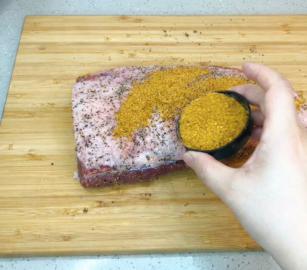 木製カッティングボードの上の大きな生牛ブリスケットに、黒い小さな器から茶褐色のバーベキューシーズニングを振りかけている様子。