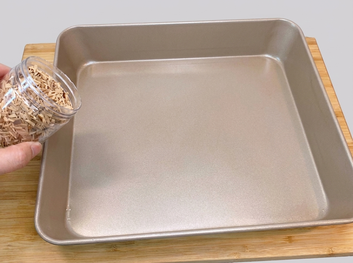 プラスチック容器から金色の四角いベーキングパンの底へ、ピーチウッドチップを均一に注ぎ入れている様子。