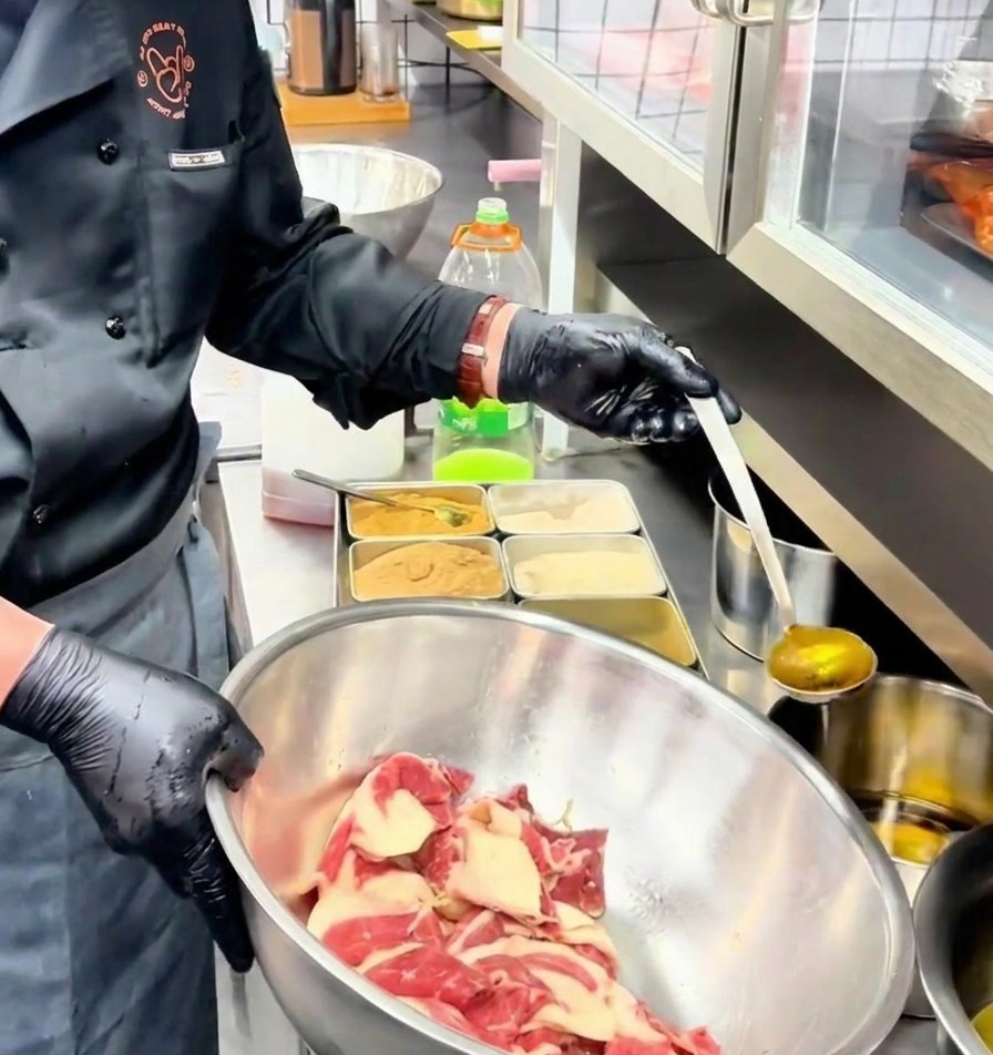 ステンレスボウルの中の味付けされた牛肉に、レードルから黄金色の香味油が注がれている様子。