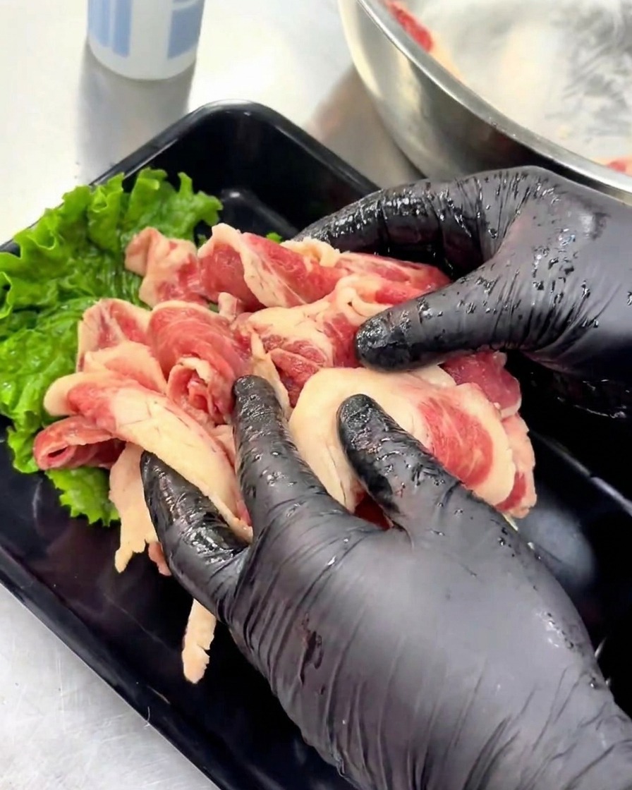 新鮮なグリーンレタスを敷いた黒いトレイの上に、味付けされた生の薄切り牛ブリスケを丁寧に並べている手元の様子。