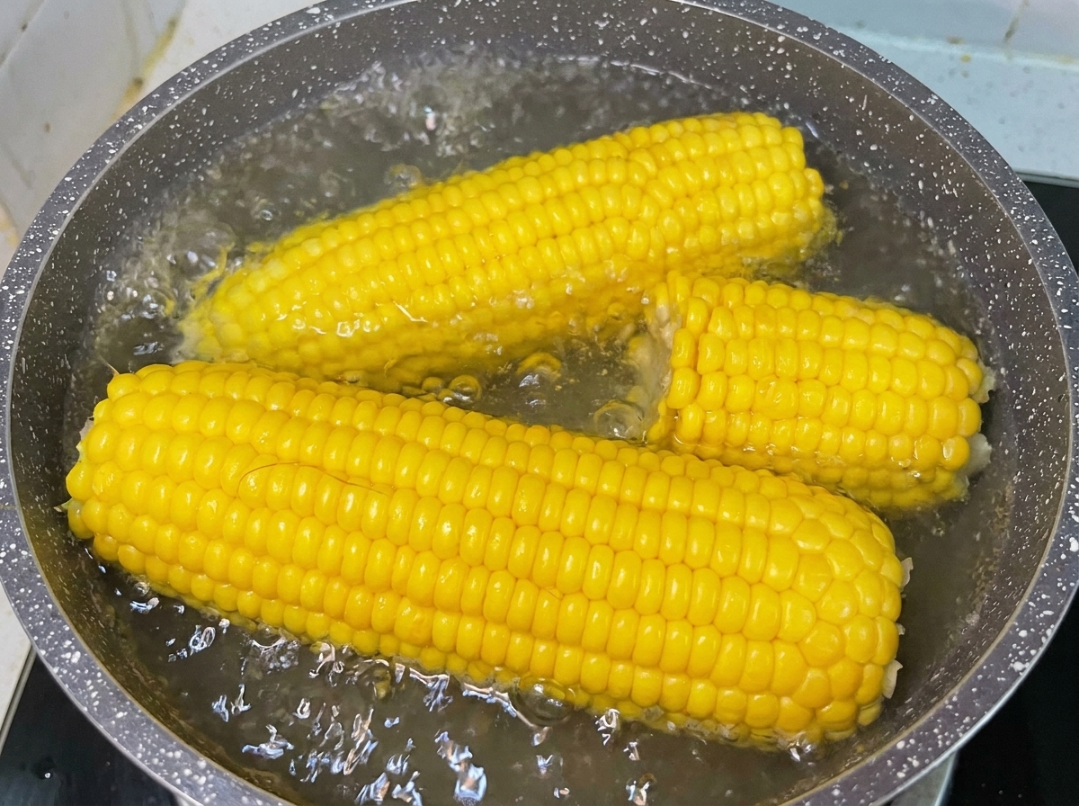 コンロの上の鍋で、お湯がぷつぷつと沸騰する中、黄色いスイートコーンを茹でている様子。
