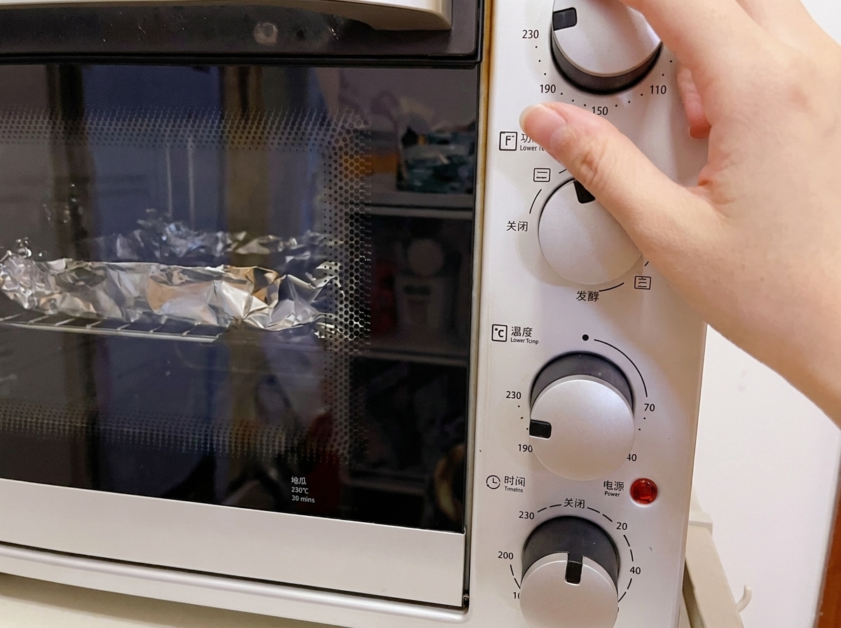 卓上型の白い電気オーブンの温度ダイヤルを230℃に合わせている手のクローズアップ。庫内にはホイルに包まれた食材が見える。
