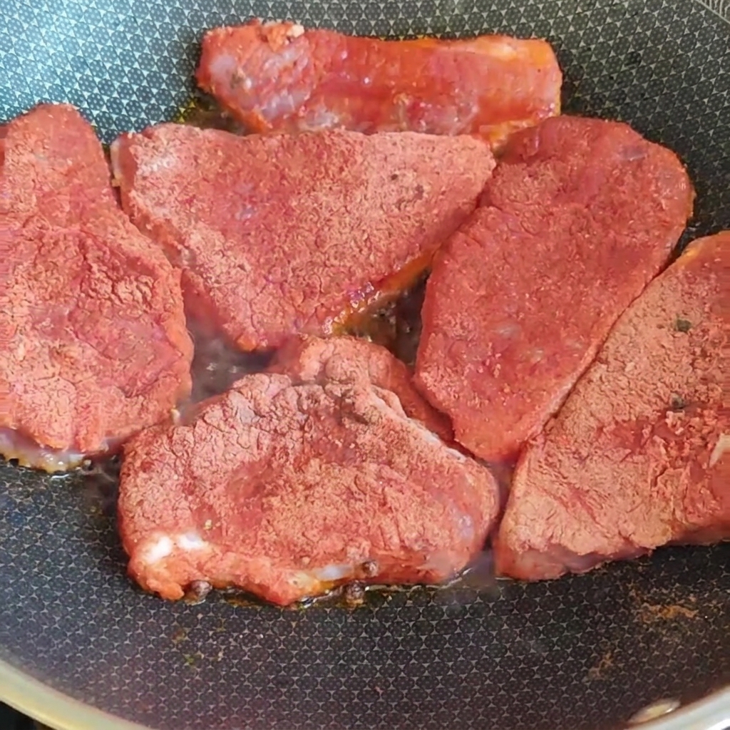 赤いスパイスミックスを全面にまぶした厚切りの豚肉を、コンロの上の熱いフライパンで香ばしく焼いている様子。