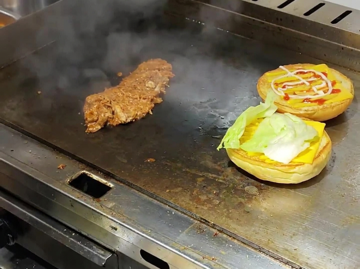 湯気が上がる鉄板の上で、軽く焼いたバーガーバンズにチーズとレタスを重ね、その横に温かいプルドポークが用意されている様子。