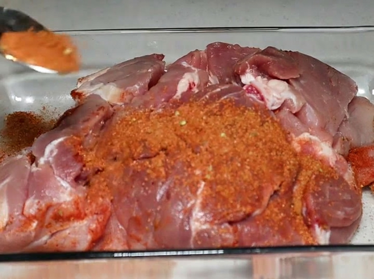 耐熱ガラスの容器に入った生の豚肩肉ブロックに、赤茶色のスパイスをスプーンでふりかけている様子。