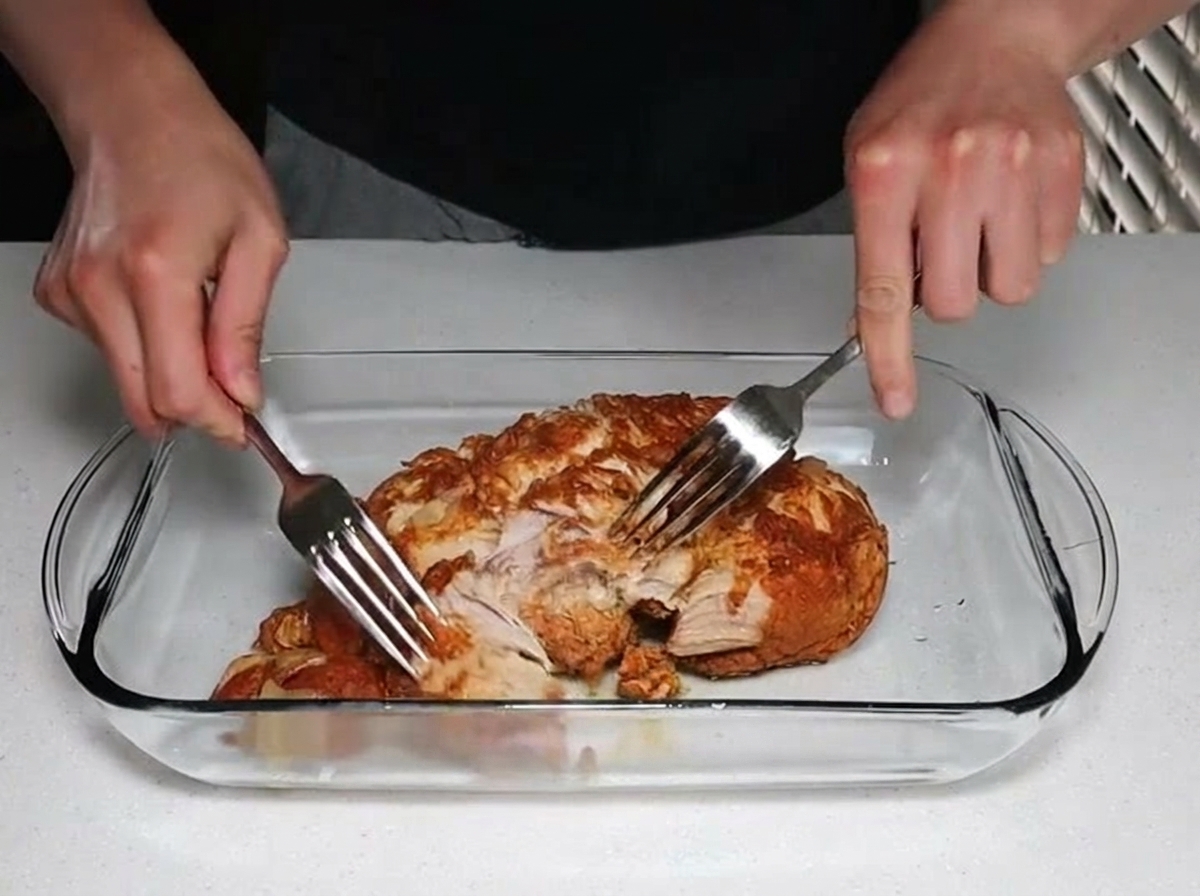 ガラスの耐熱容器の中で、調理された柔らかい豚肉を2本のフォークを使って引き裂き、ほぐしている両手。