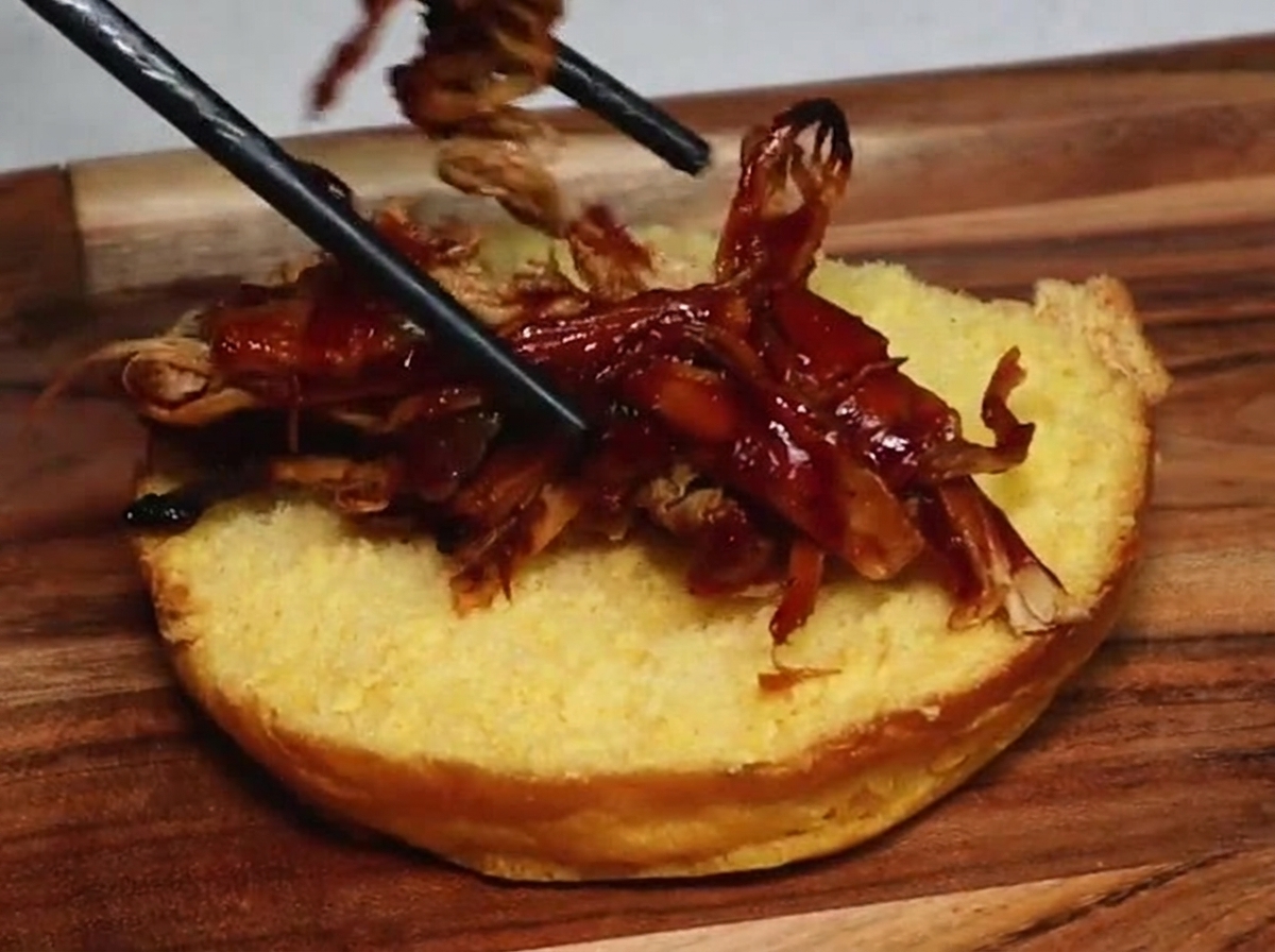こんがりと焼いたバーガーバンズの下半分に、お箸を使ってバーベキューソースが絡んだジューシーなプルドポークを乗せている様子。