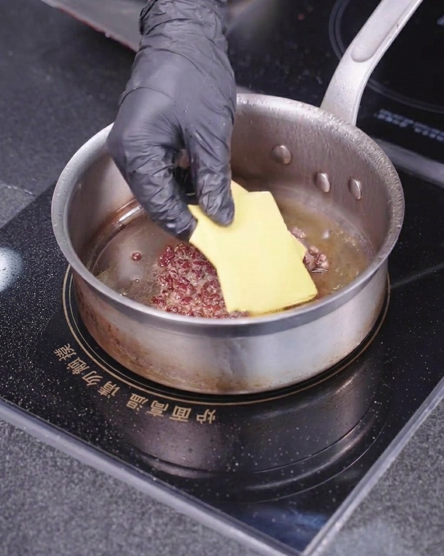 熱いスキレットの中で焼かれている肉パティの上に、手袋をはめた手で四角い黄色いチーズをのせている様子。