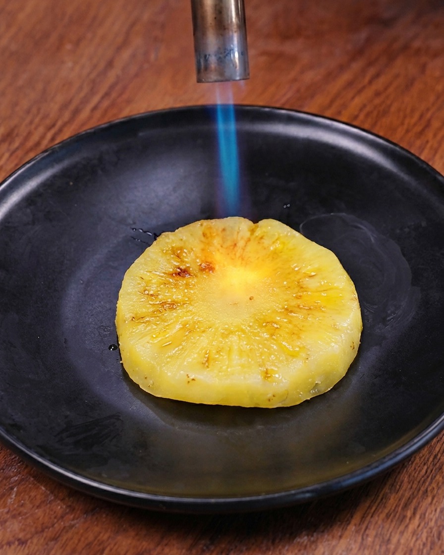 黒い皿の上で、新鮮な輪切りパイナップルの表面をキッチンバーナーの青い炎で炙り、焼き目をつけている様子。