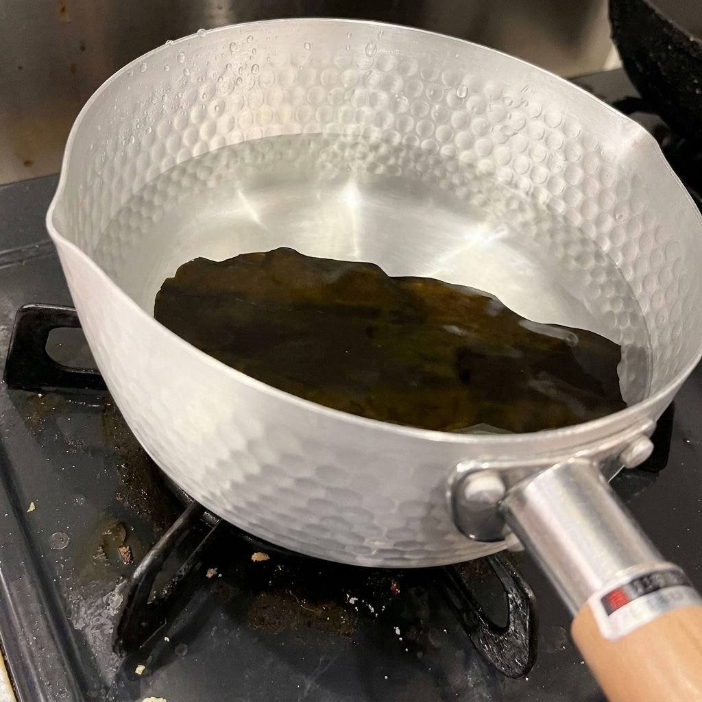 澄んだ冷たい水が入ったアルミ鍋の底に、乾燥した昆布が一枚沈んでいる様子。