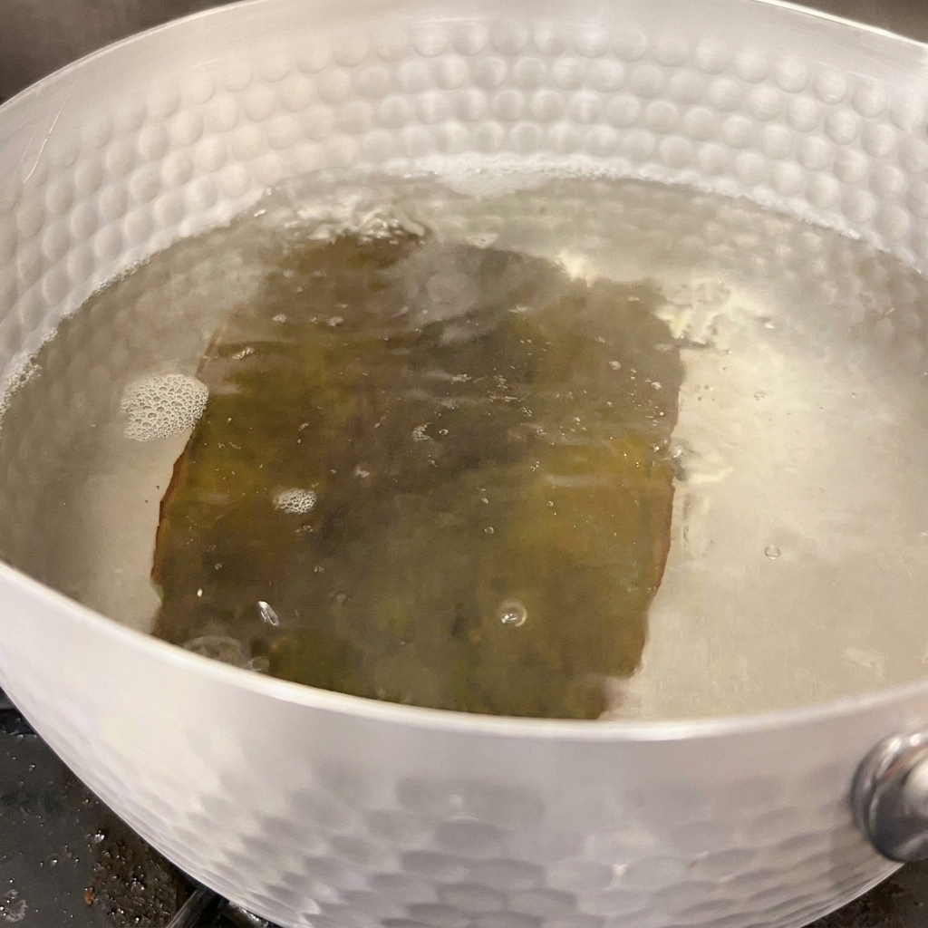 コンロの上の銀色の鍋の中で、戻った昆布の周りで水が優しく泡立っている様子。