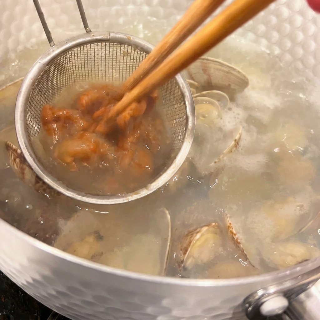 小さな味噌漉しの中の茶色い味噌を、箸を使って熱いあさりのスープに混ぜながら溶かしている様子。