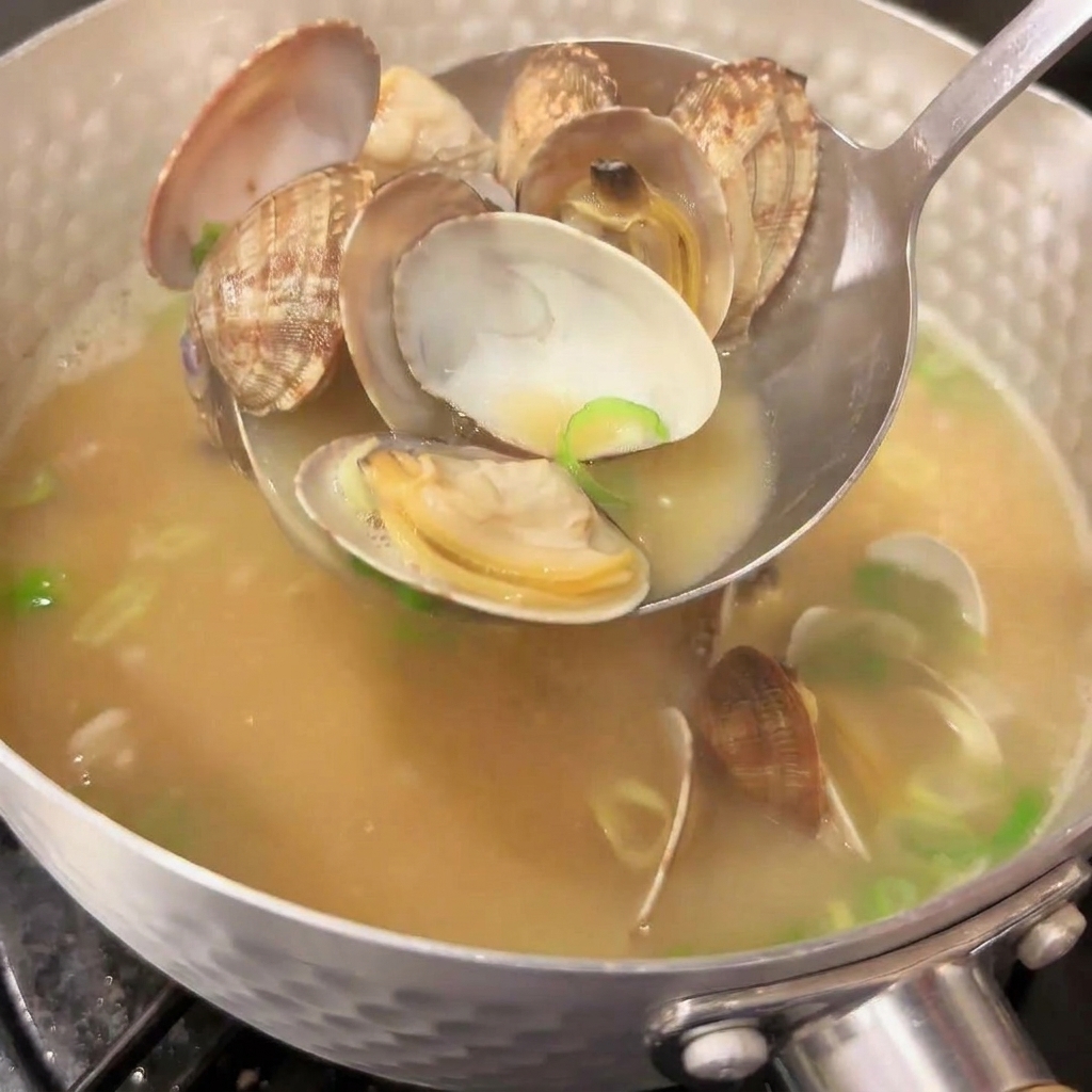 湯気が立つ熱々のみそ汁の鍋から、口の開いたあさりをすくい上げる金属製のお玉。スープには鮮やかな緑の刻み青ねぎが散りばめられている。