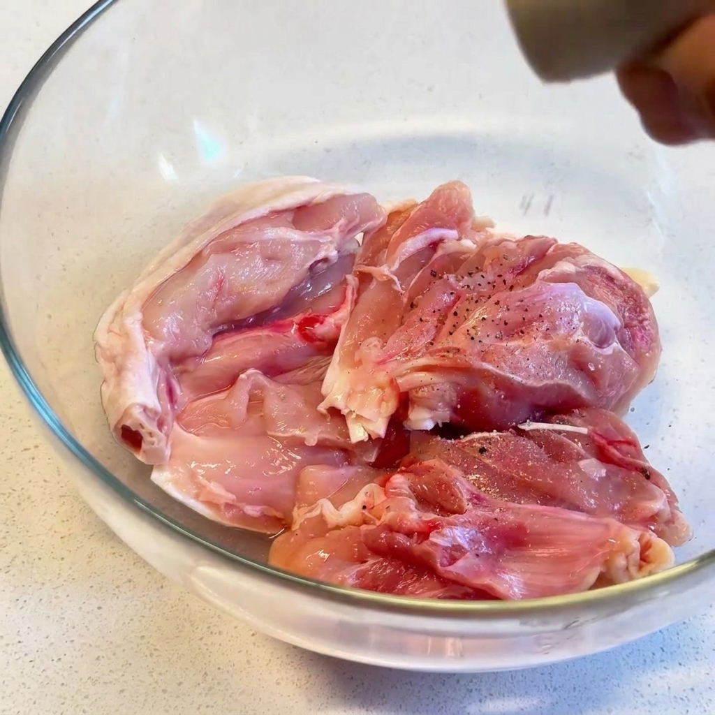 透明なガラスボウルに入った生鶏もも肉に、ペッパーミルで挽きたての黒こしょうをかけている様子。