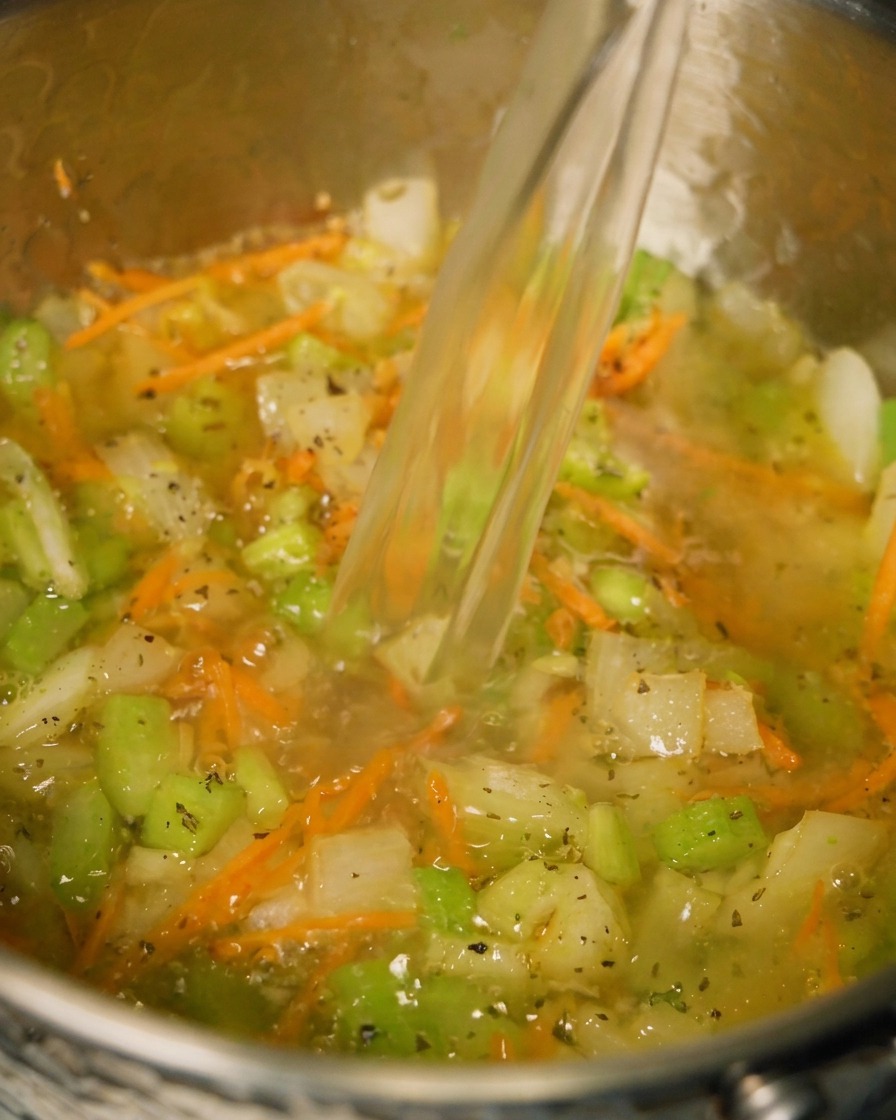炒めた野菜とハーブが入った鍋に、チキンブイヨンを注ぎ入れている様子。