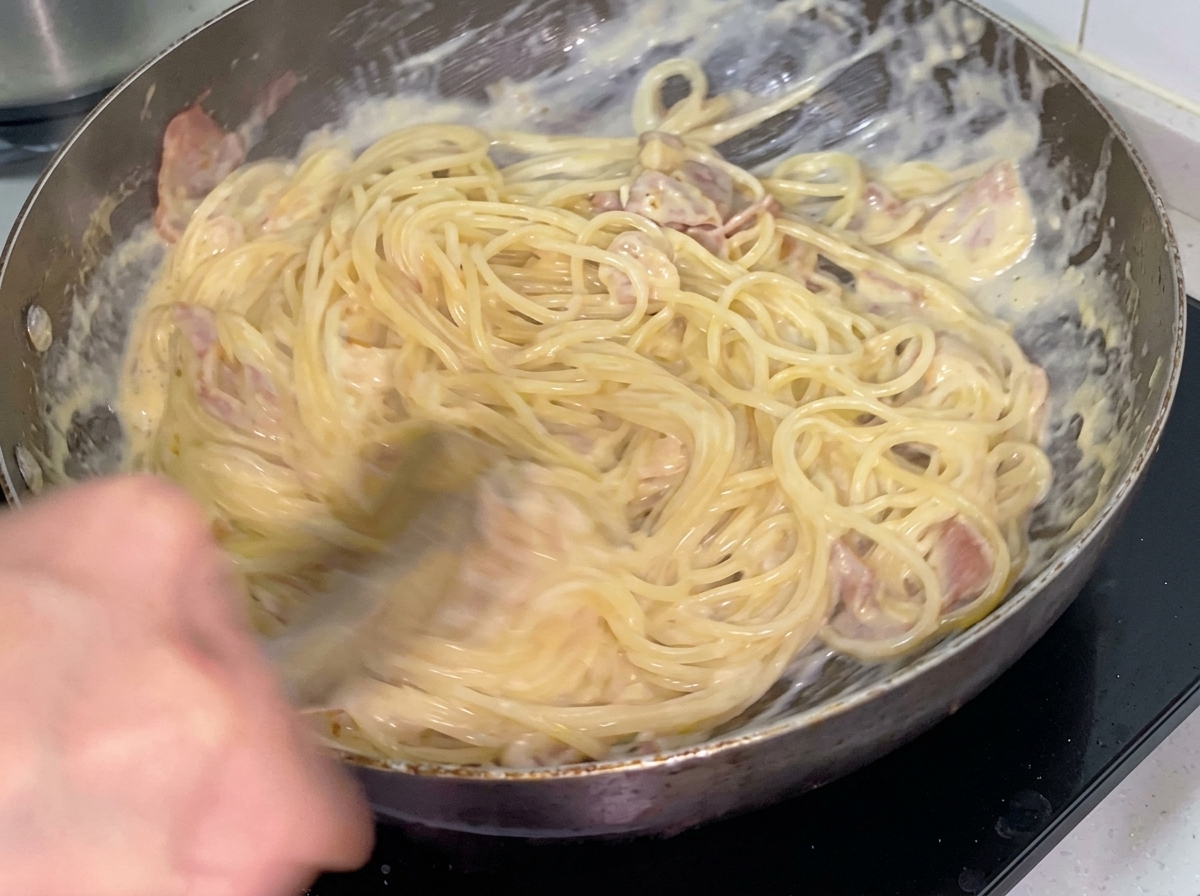 金属製のトングを使って、クリーミーなスパゲッティをベーコンや鶏肉と一緒に混ぜ合わせている様子。