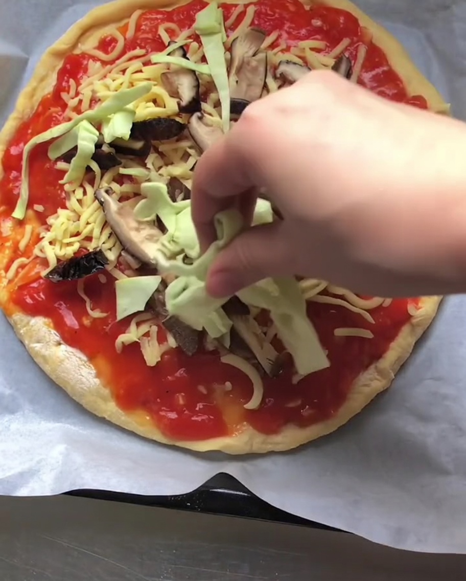 トマトソースの上にチーズとスライスした椎茸がのったピザに、生の千切りキャベツを散らしている手。
