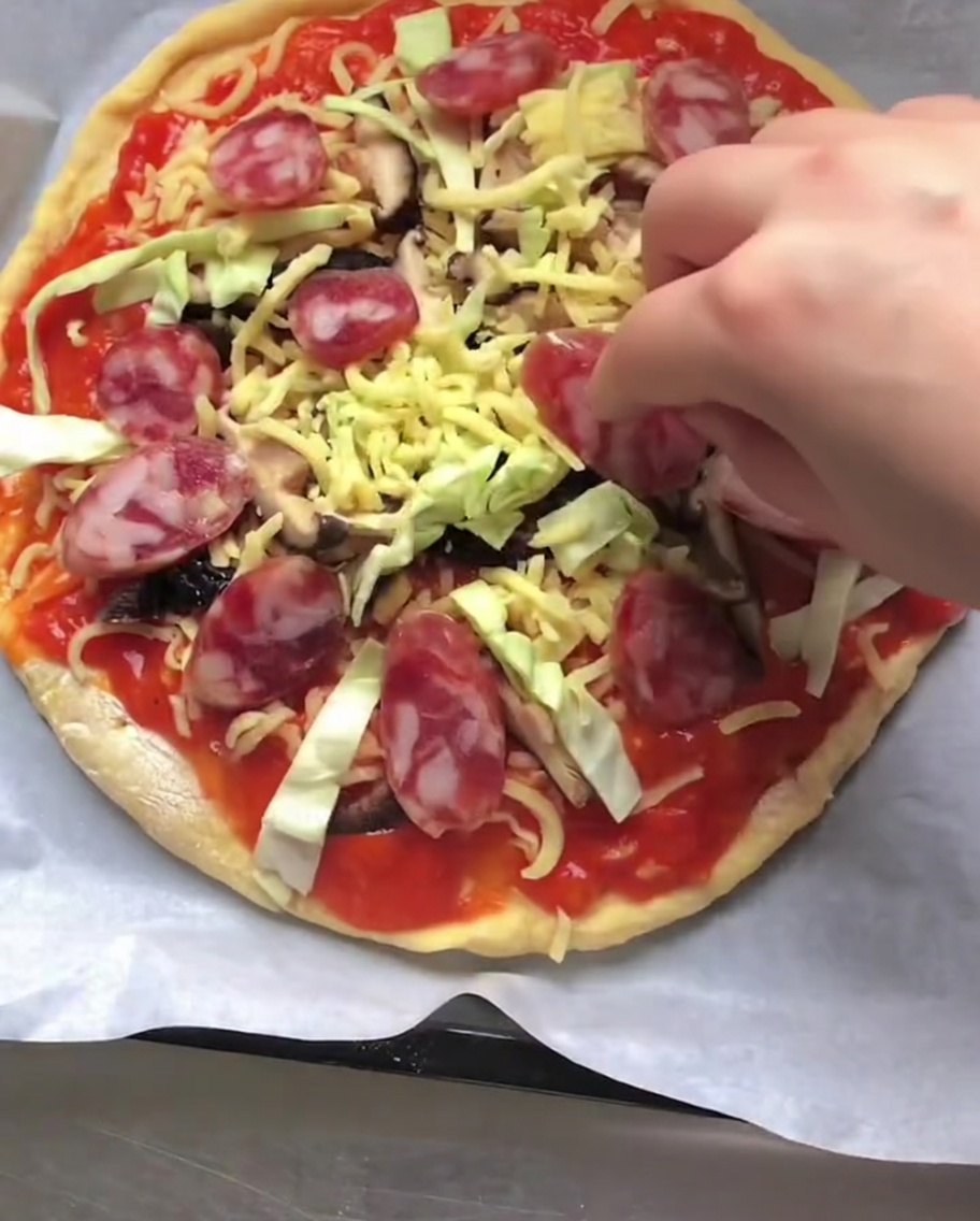 ソース、チーズ、野菜がのった焼成前のピザに、赤いイタリアンソーセージのスライスを並べている手。