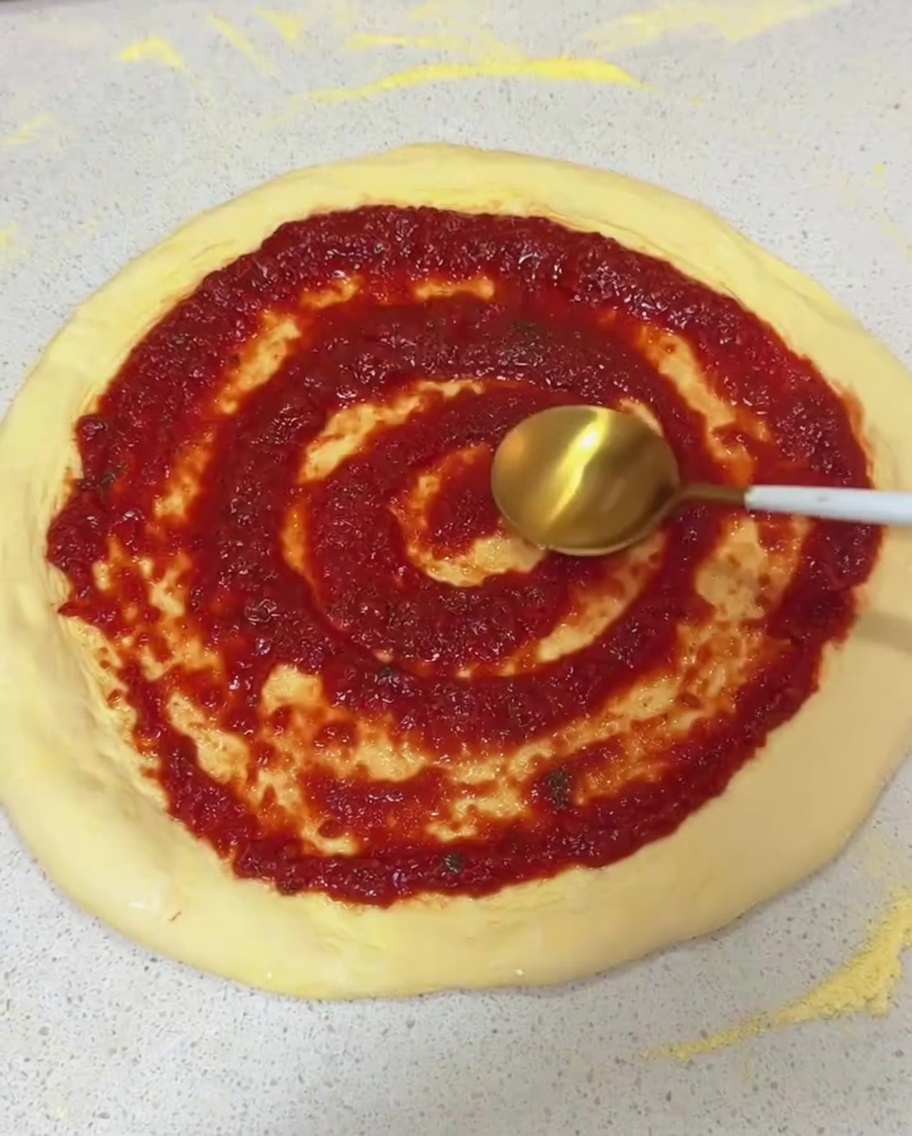 伸ばした生のピザ生地に、スプーンの背を使って円を描くように赤いトマトソースを塗っている様子。