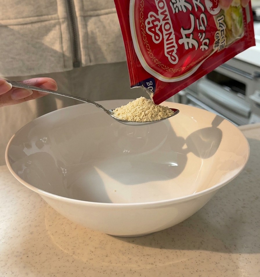 大きな白い丼に、スプーンで鶏ガラスープの素を入れている様子。