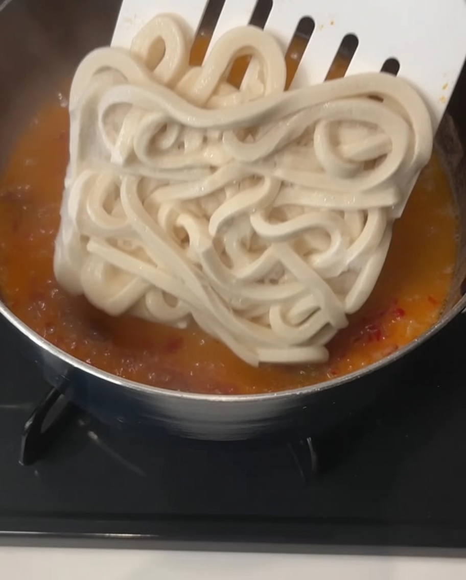 沸騰しかけているオレンジ色のスープに、冷凍うどんの塊を投入している様子。