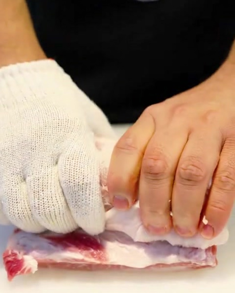 手袋をした手で、生の豚バラ肉をタコ糸でしっかりと縛っている様子。