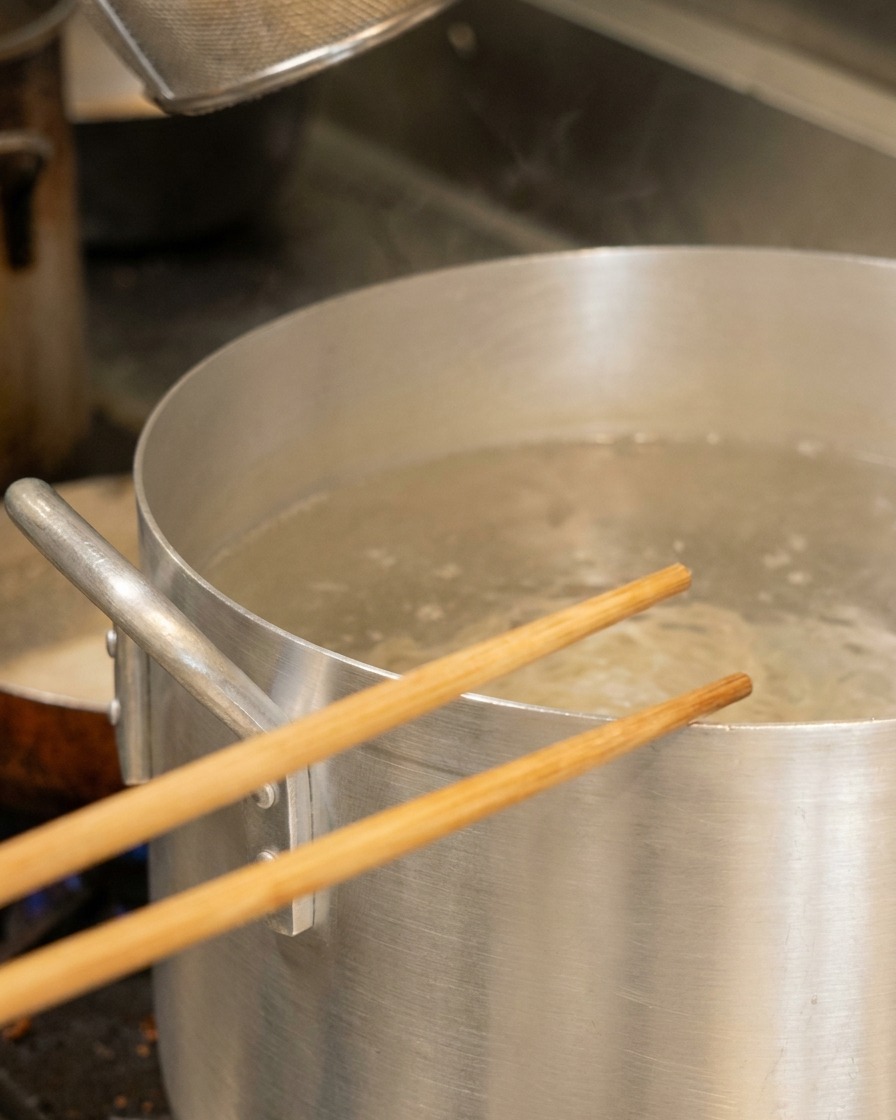 麺用のざるが入った沸騰した大きなアルミ鍋の上に、木製の箸が置かれている。