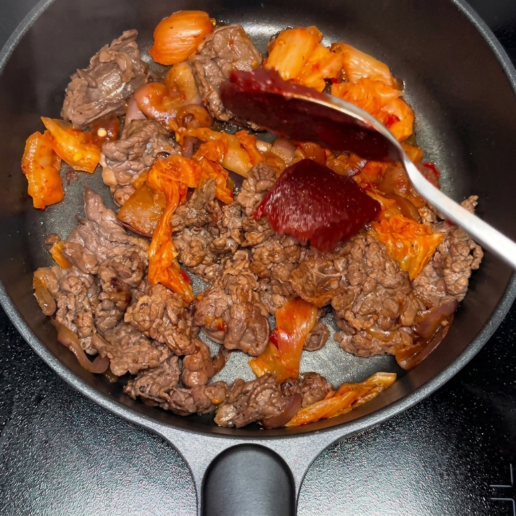 炒めた牛肉とキムチの混合物にコチュジャンと粉唐辛子を加えている様子。