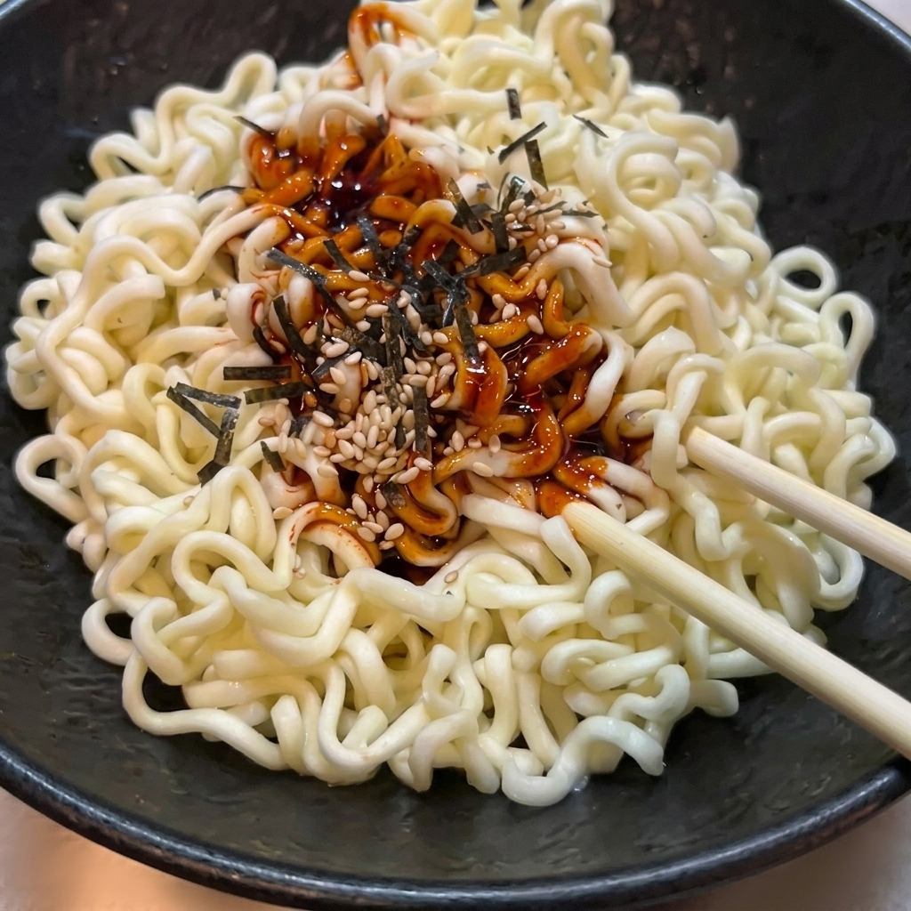 黒いボウルに入った茹でたウェーブ麺に、赤いソース、ゴマ、海苔を混ぜ合わせている様子。