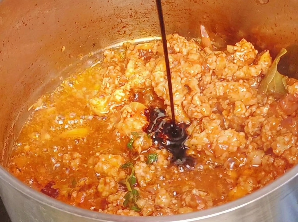赤みがかったミートソースが入った鍋に醤油を注いでいる様子。