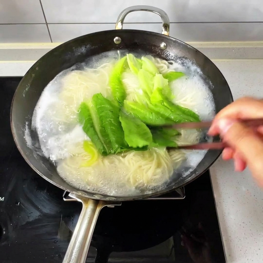 沸騰したお湯の中の麺と青菜を、木製の箸で混ぜている様子。