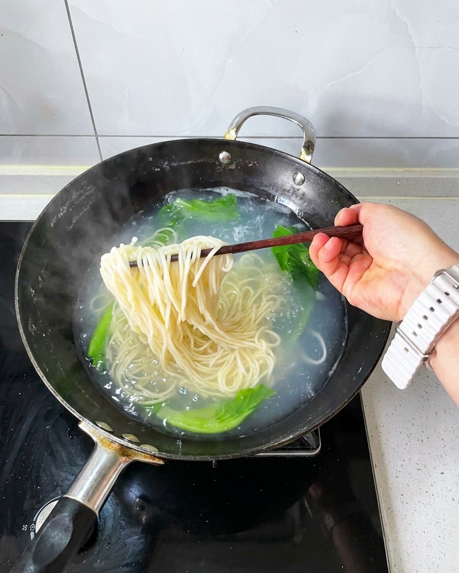木製の箸で、茹で上がった麺と青菜をお湯から引き上げている様子。