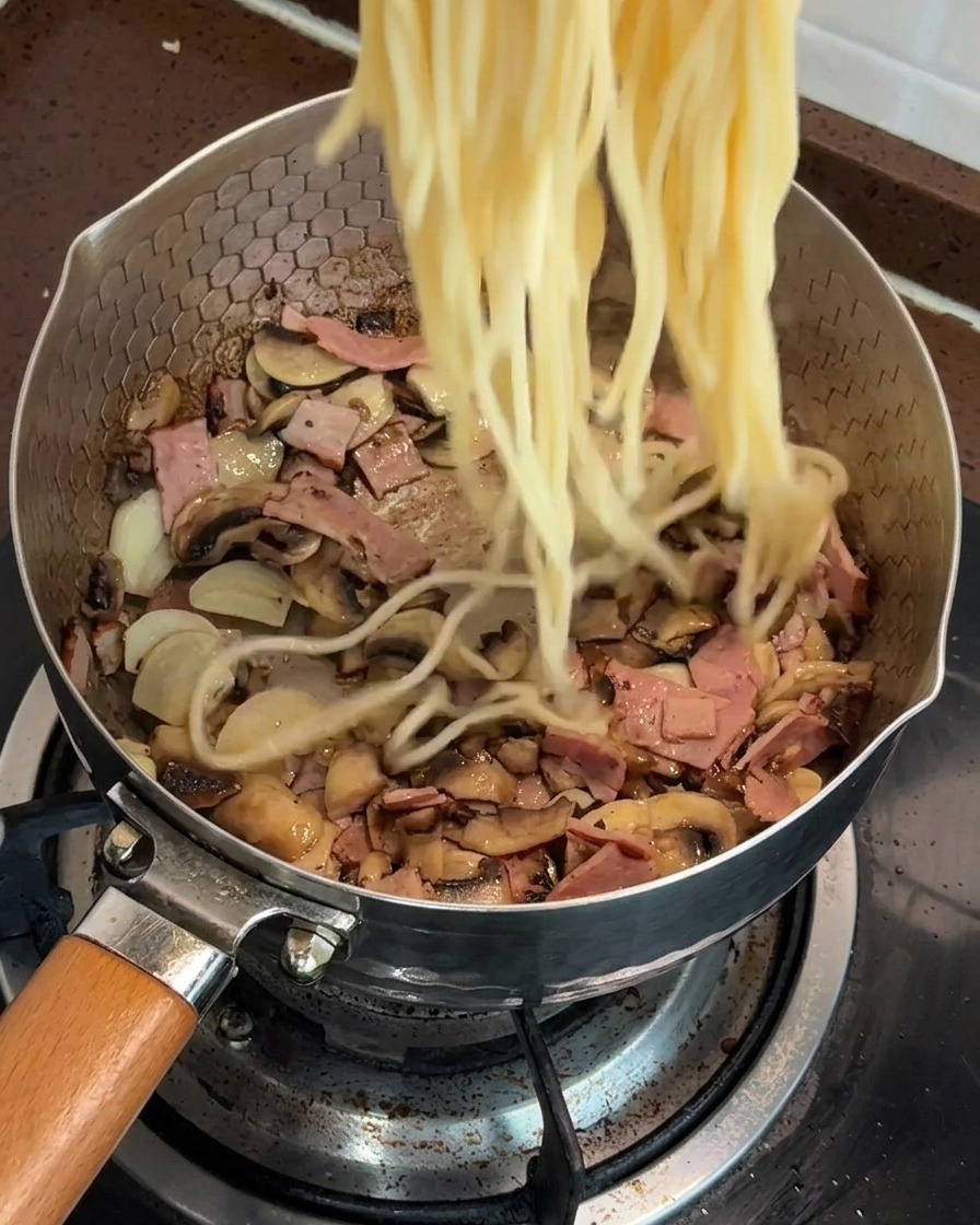 炒めたマッシュルームとベーコンが入ったフライパンに、茹でたスパゲッティを加えている様子。