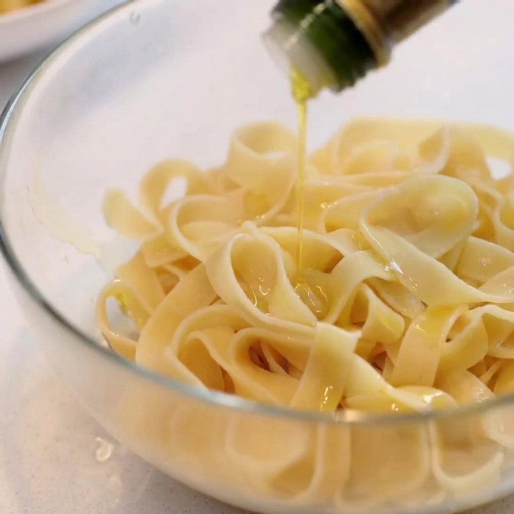 ガラスのボウルに入った茹でた平打ちパスタにオリーブオイルを回しかける様子。