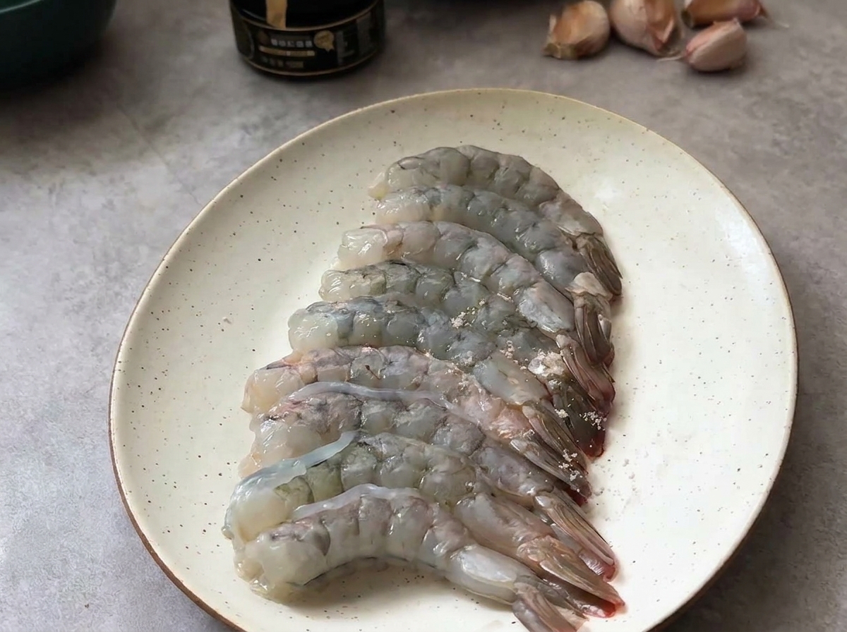 皿に並べた生のむきエビに、塩と黒こしょうで軽く下味をつけている様子。