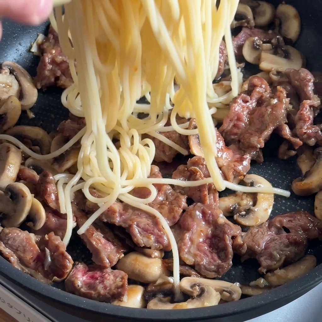 牛肉と炒めたきのこが入ったフライパンに、茹でたてのパスタを加える様子。