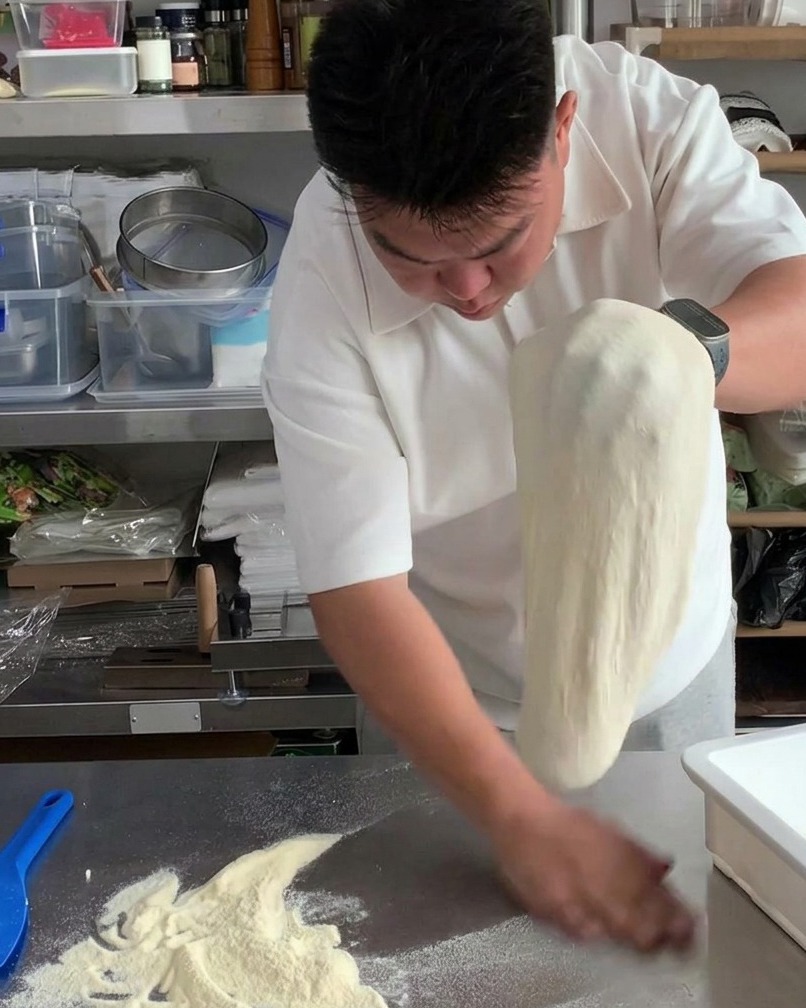伸ばしたピザ生地を両手で交互に入れ替えながら、表面の余分な打ち粉を落としている様子。