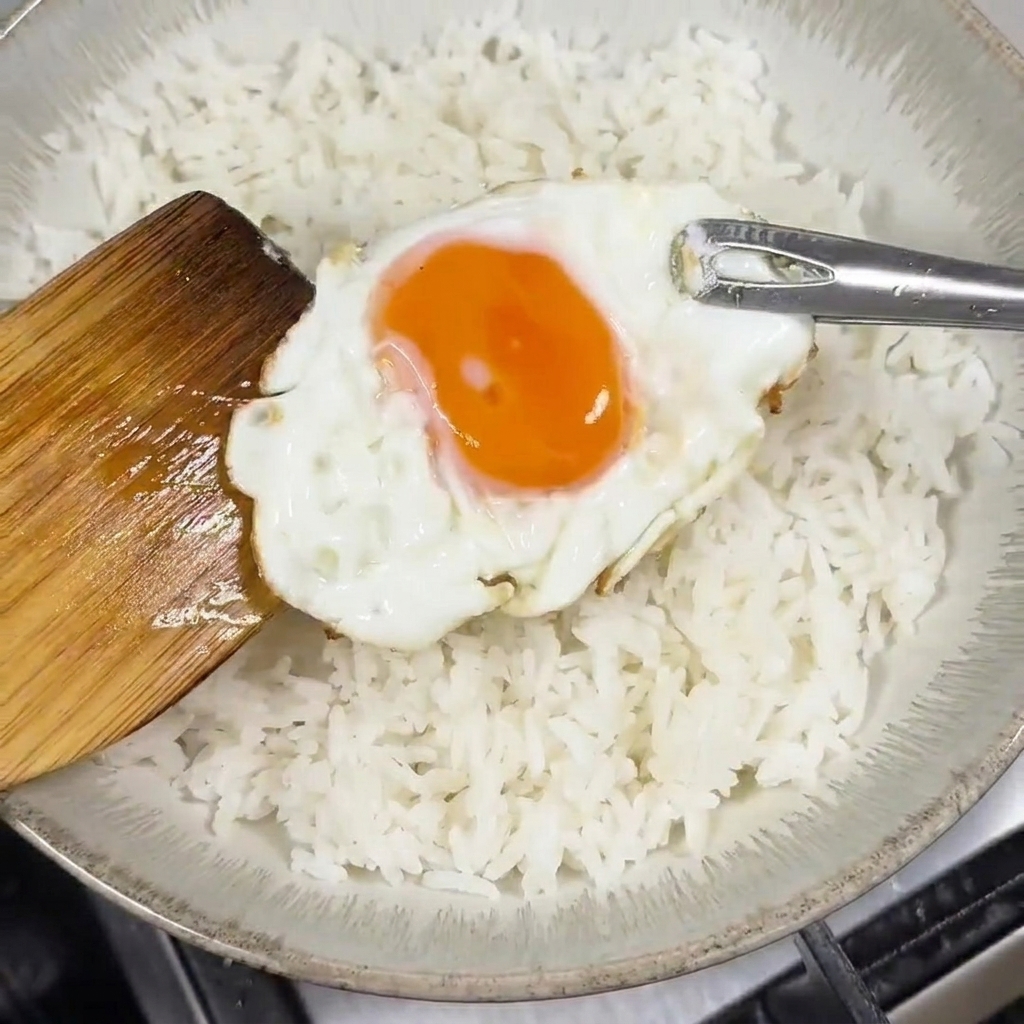 白いご飯を盛った丼の真ん中に、半熟の目玉焼きを木べらでそっとのせる様子。