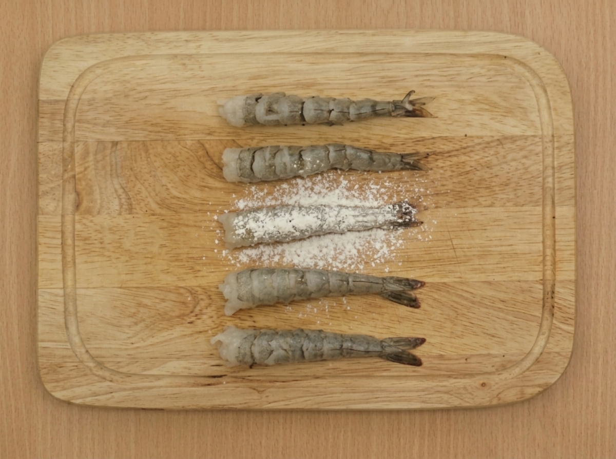 木製のボードに並べられた下処理済みの海老に、粉ふるいで薄力粉をまぶしている様子。