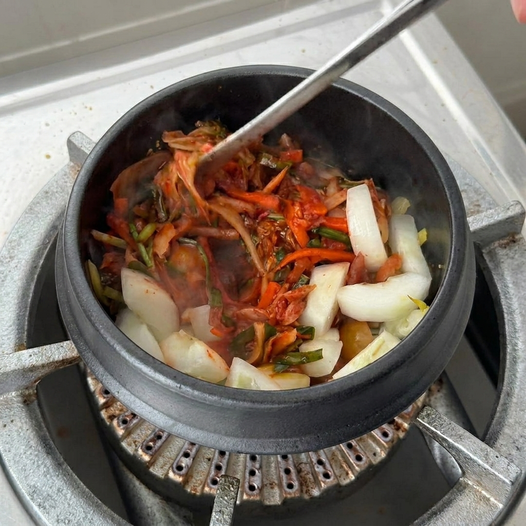 大きなカップに入ったキムチのスープベースを、炒めたキムチと玉ねぎが入った土鍋に注ぎ入れている様子。