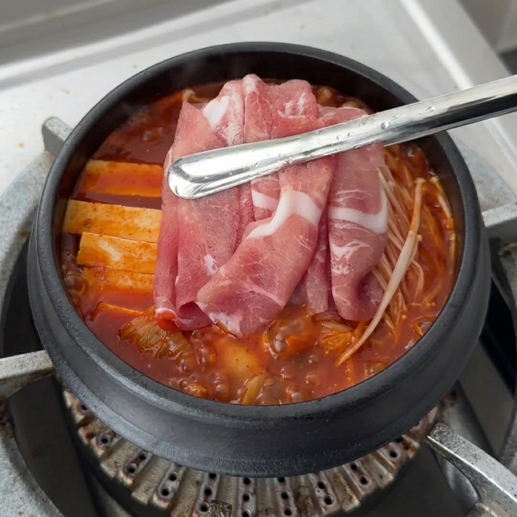 生の豚肉のスライスを、煮込んでいるキムチチゲの具材の上に並べている様子。